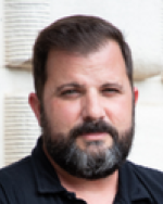 A photo of Richard from the shoulders up while he is standing outside of a building and wearing a black polo shirt.