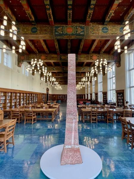 A grand library reading room with long wooden tables and tall bookshelves is centered around a tall pink marble obelisk, beneath decorative ceiling beams and large circular chandeliers.
