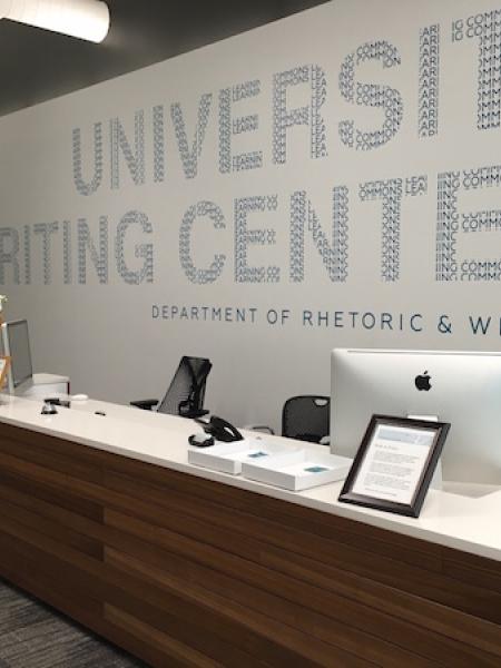 A modern reception desk area with computers and signage on the wall reading “University Writing Center, Department of Rhetoric & Writing.”