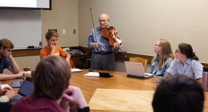 An instructor stands at a table holding a violin while demonstrating to a group of seated students in a classroom setting.